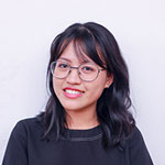 A smiling young woman with dark wavy hair and round glasses wearing a black top against a white background.