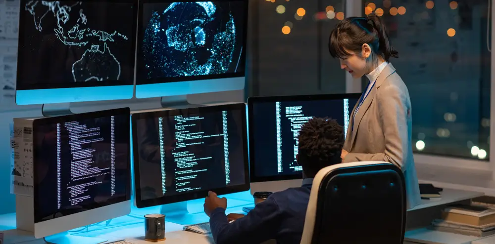 Two IT professionals working at a multi-monitor workstation displaying code and global data maps at night.