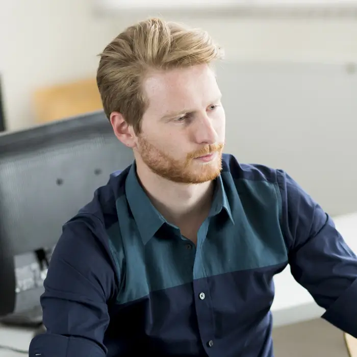 A young man with a beard and light brown hair wearing a blue button-down shirt, looking thoughtfully to the side.