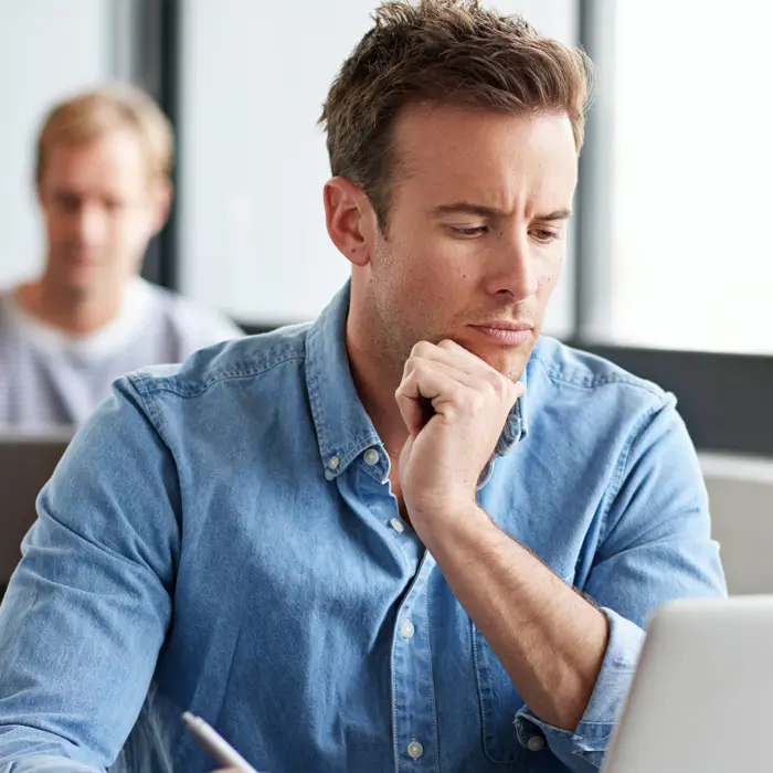 A man in a blue denim shirt looking thoughtfully at a laptop screen in a bright office setting.