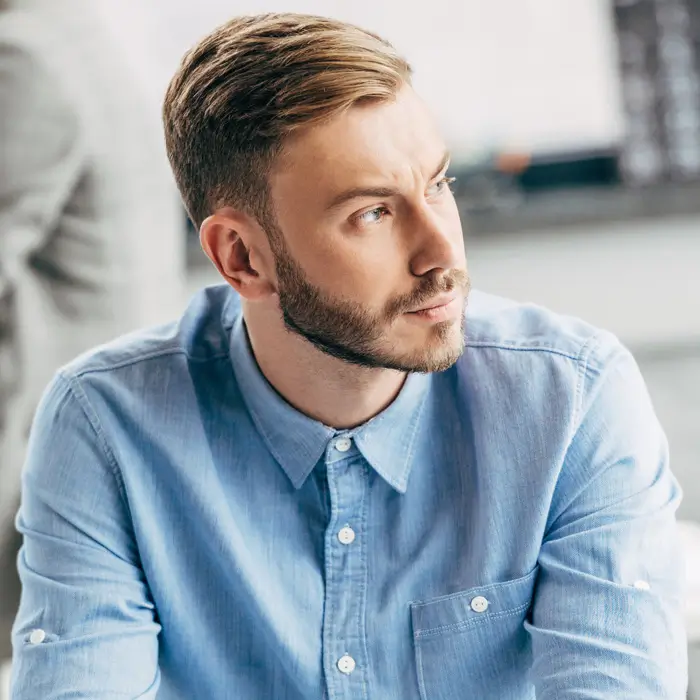 A man with a beard and styled hair wearing a blue button-down shirt.